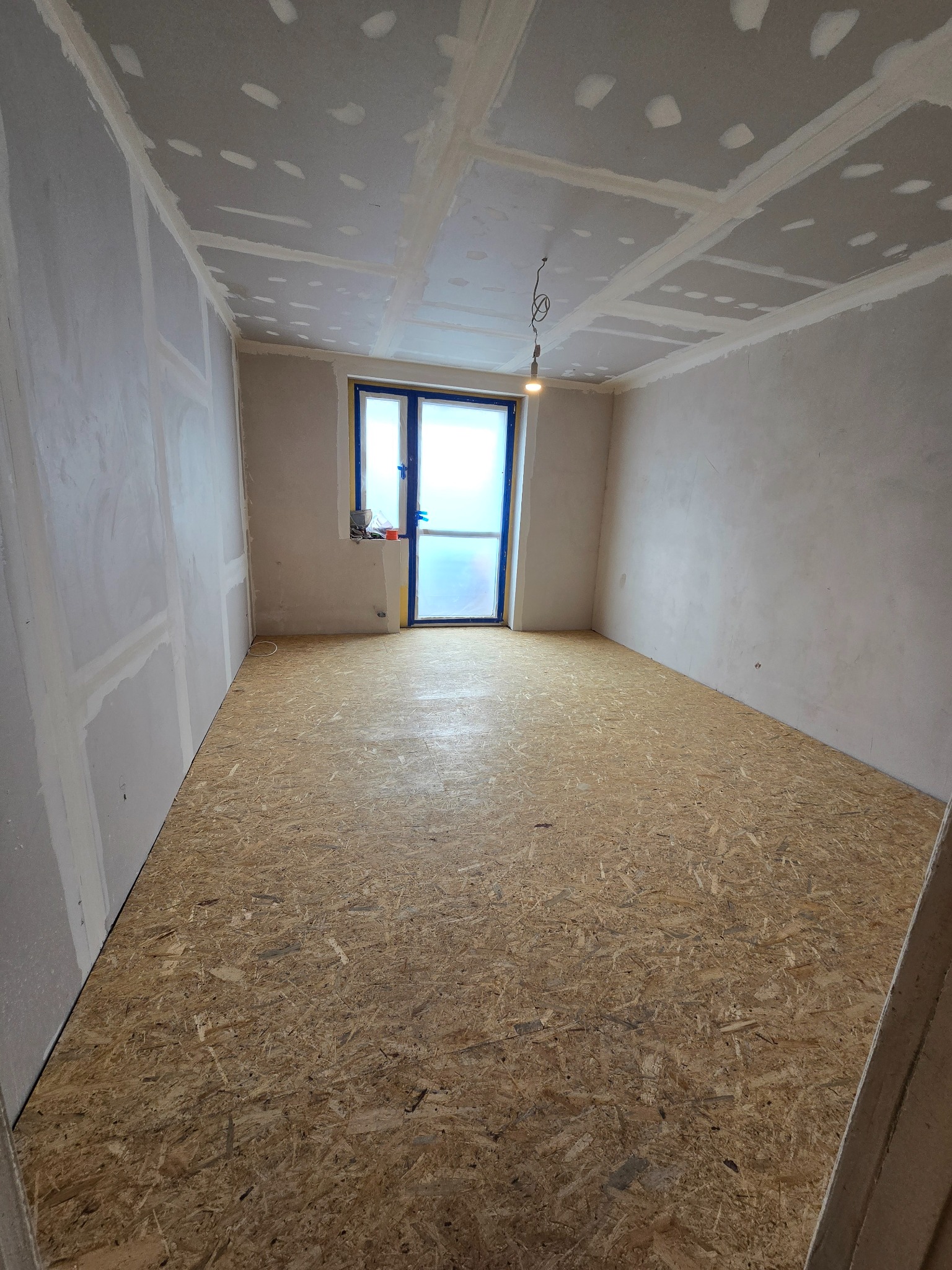 Pokój w trakcie remontu: ściany przygotowane pod gładzie, sufit z płyt gipsowych, podłoga wyłożona płytami OSB, okno zabezpieczone folią malarską.