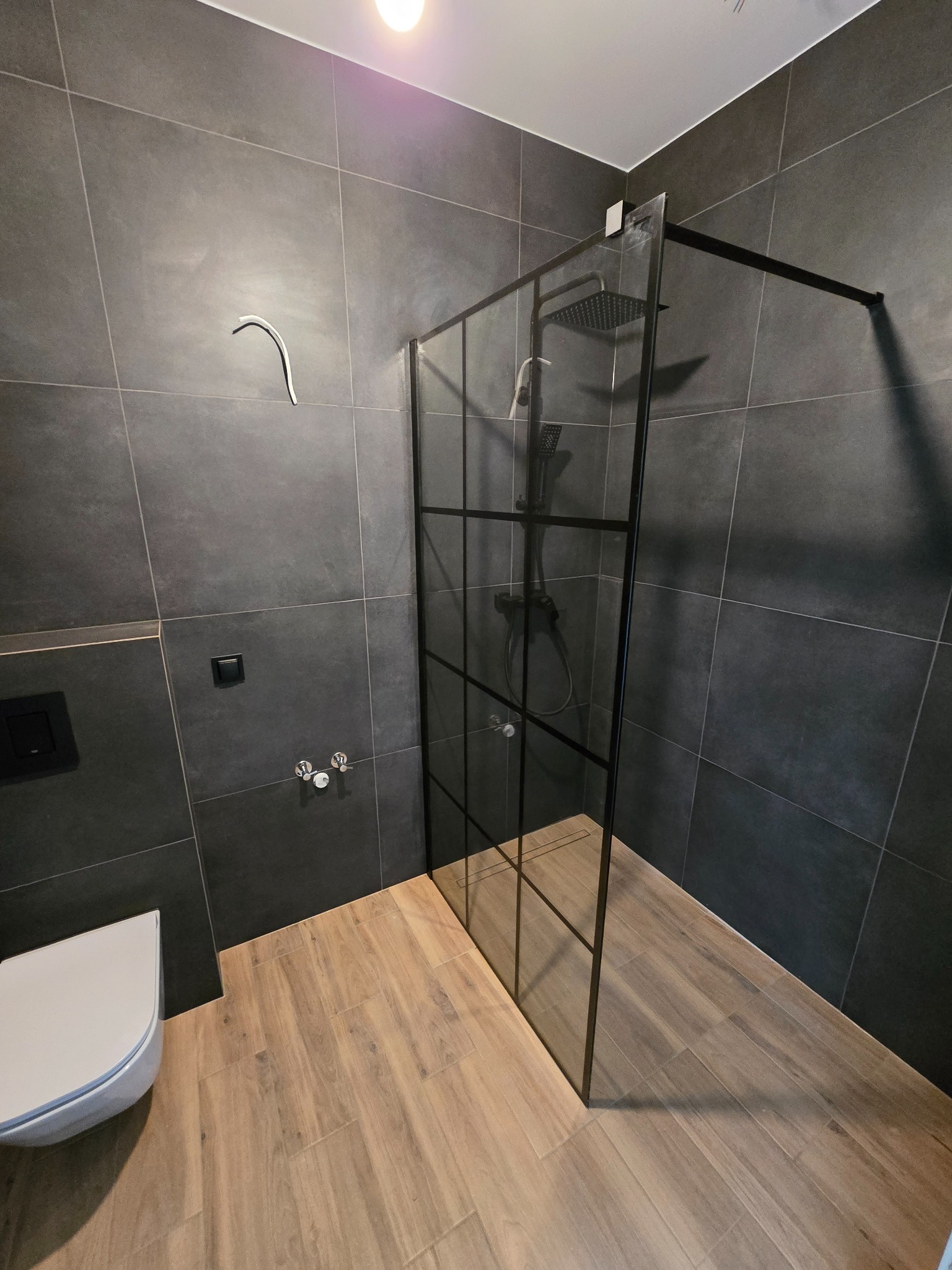 Nowoczesna łazienka z kabiną prysznicową typu walk-in z czarnymi profilami, szare płytki na ścianach i drewnopodobna podłoga. Minimalistyczny design.