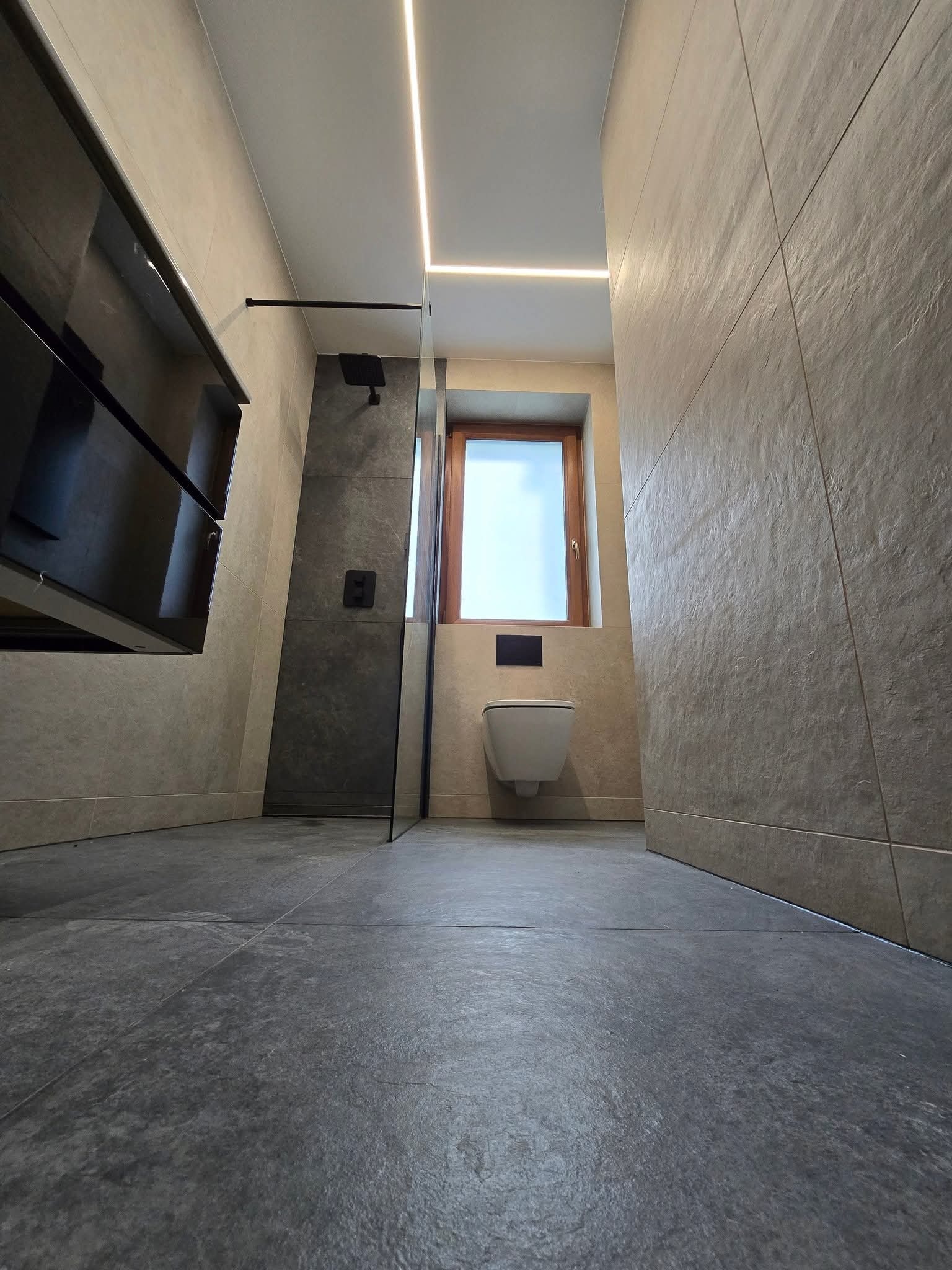 Nowoczesna łazienka z szarą podłogą, beżowymi płytkami na ścianach i podświetleniem LED. Kabina prysznicowa ze szklaną ścianką i wisząca toaleta.