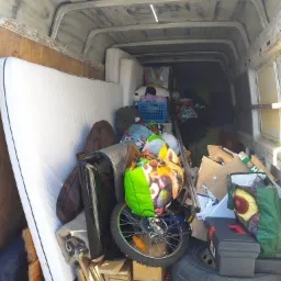 Wnętrze furgonetki wypełnionej r&oacute;żnorodnymi przedmiotami podczas załadunku, widać materac, rower, pudła, torby z zakupami, oponę i skrzynkę narzędziową.