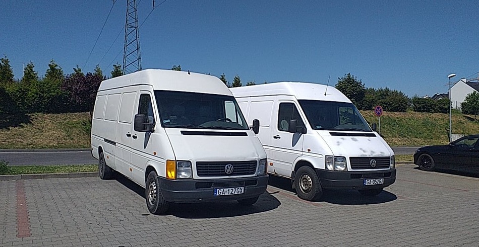 Dwa białe busy Volkswagen LT na parkingu z kostki brukowej, w tle zieleń i słup wysokiego napięcia.