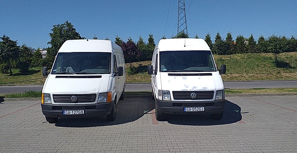 Dwa białe busy Volkswagen LT zaparkowane obok siebie na brukowanym parkingu w słoneczny dzień, widoczne numery rejestracyjne GA 127GX i GA 052GJ.