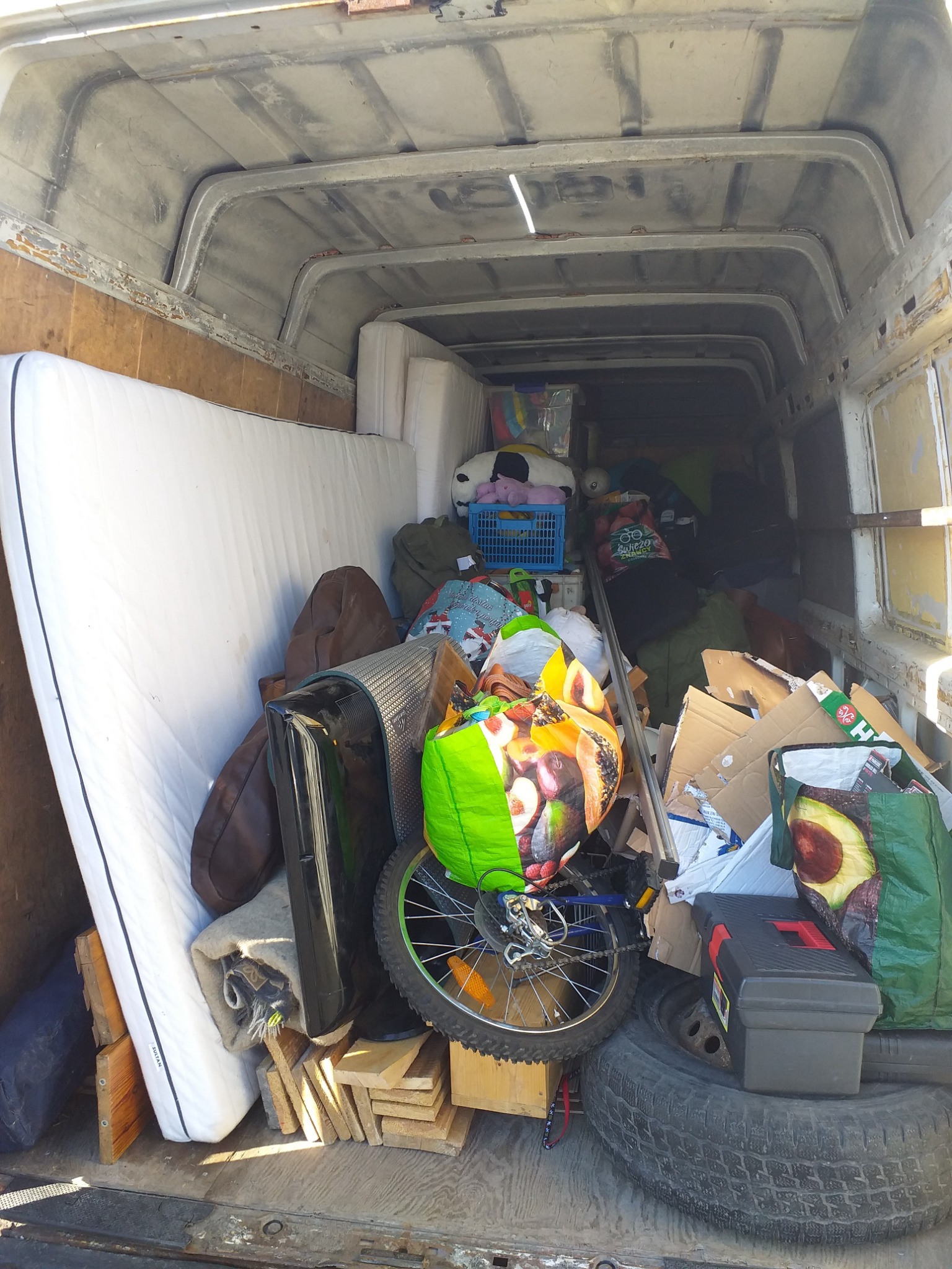 Wnętrze furgonetki wypełnionej różnorodnymi przedmiotami podczas załadunku, widać materac, rower, pudła, torby z zakupami, oponę i skrzynkę narzędziową.