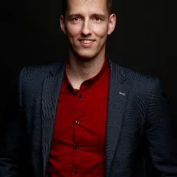 Portret uśmiechniętego mężczyzny w ciemnogranatowej marynarce i czerwonej koszuli na ciemnym tle, z przypinką w kształcie agrafki w klapie marynarki.