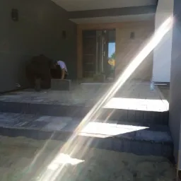 Dwóch pracowników układa szare kostki brukowe na schodach wejściowych do domu, widoczna butelka wody i piasek. Silne promienie słoneczne.