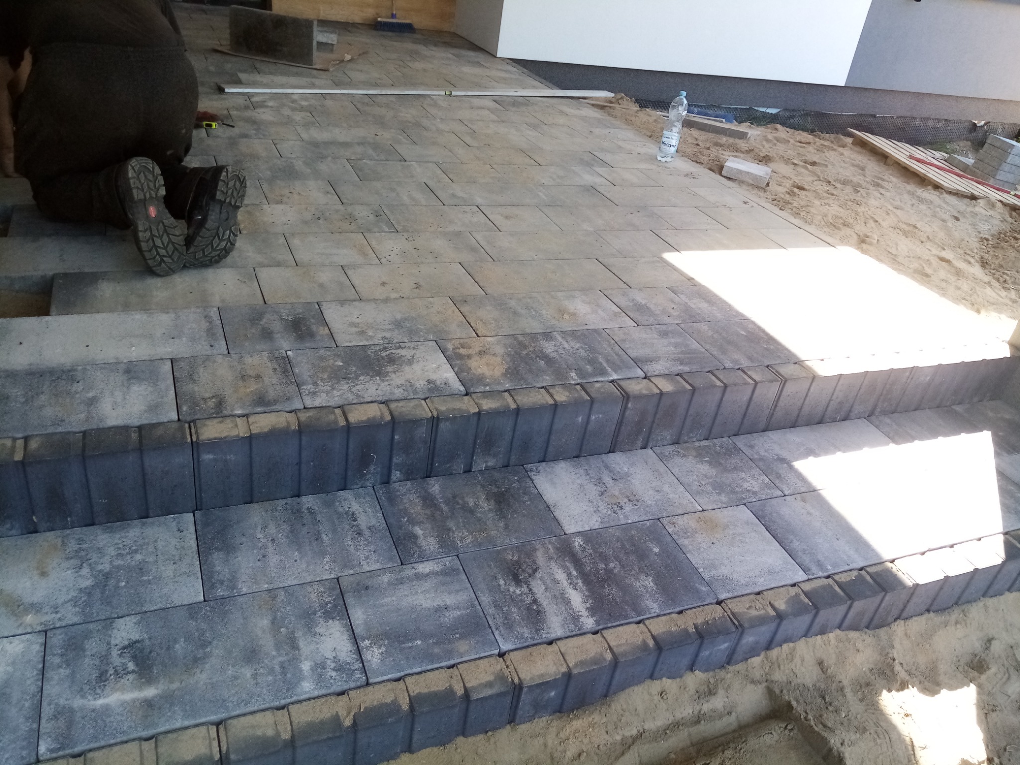 Układanie schodów z kostki brukowej w trakcie prac, widoczne buty brukarza, poziomica, piasek i butelka wody.