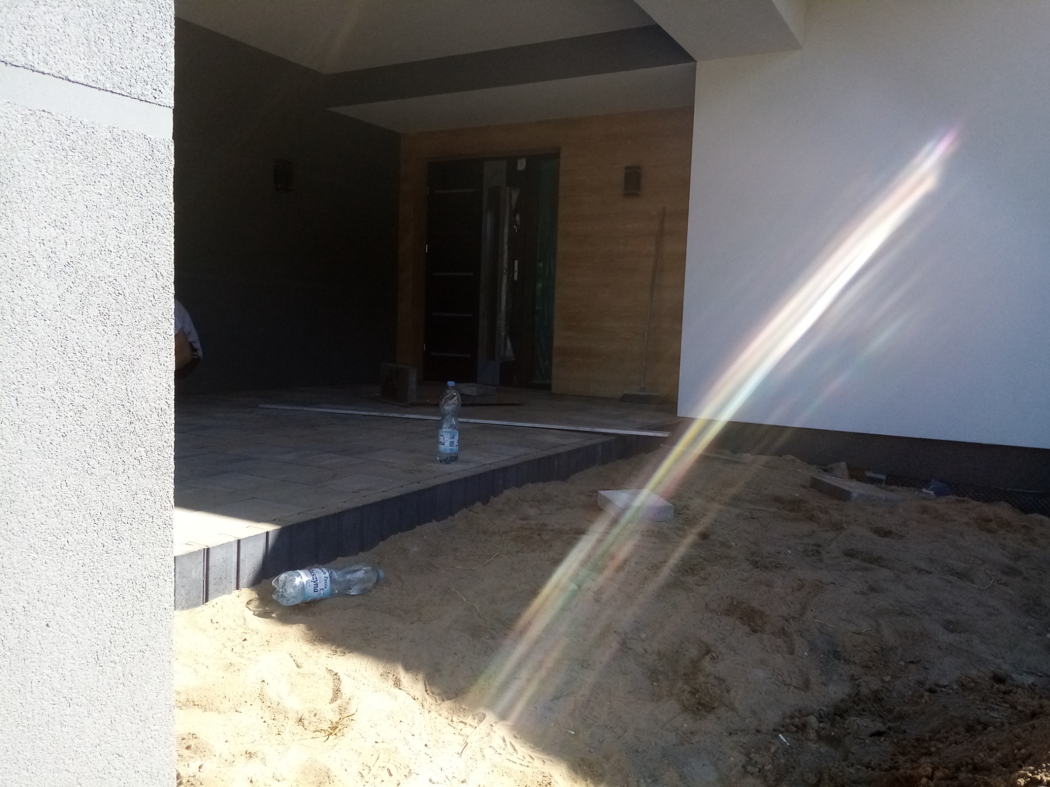 Etap przygotowania podłoża pod nawierzchnię z kostki brukowej, widoczny piasek, częściowo ułożona kostka i butelki z wodą, jasne światło słoneczne.