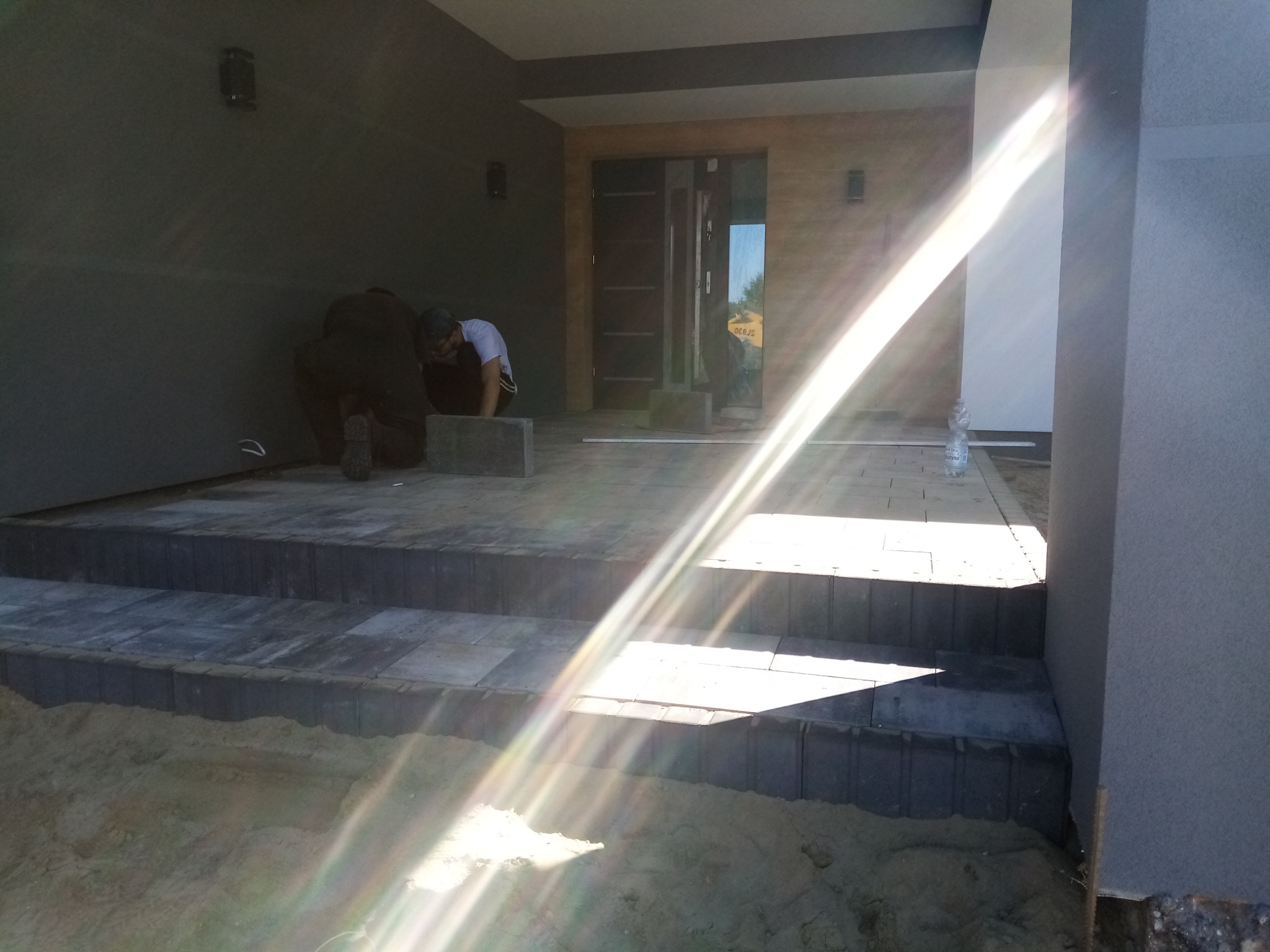 Dwóch pracowników układa szare kostki brukowe na schodach wejściowych do domu, widoczna butelka wody i piasek. Silne promienie słoneczne.