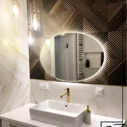 Nowoczesna łazienka z umywalką nablatową, złotą armaturą, podświetlanym lustrem owalnym na tle ściany z drewnianych paneli w geometryczny wzór oraz wiszącymi lampami ze szklanymi kloszami.
