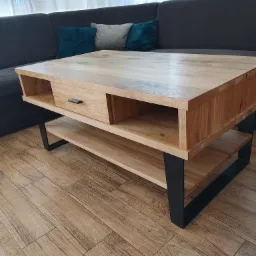 Stolik kawowy z jasnego drewna z szufladą i półką, na czarnych metalowych nogach, wykonany na zamówienie w Olszynie, widoczny na tle szarej kanapy i drewnopodobnej podłogi.