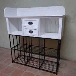 Biała komoda z dwiema szufladami i otwartymi półkami, umieszczona na czarnym metalowym stelażu z półkami, na tle beżowej ściany i terakotowej podłogi.