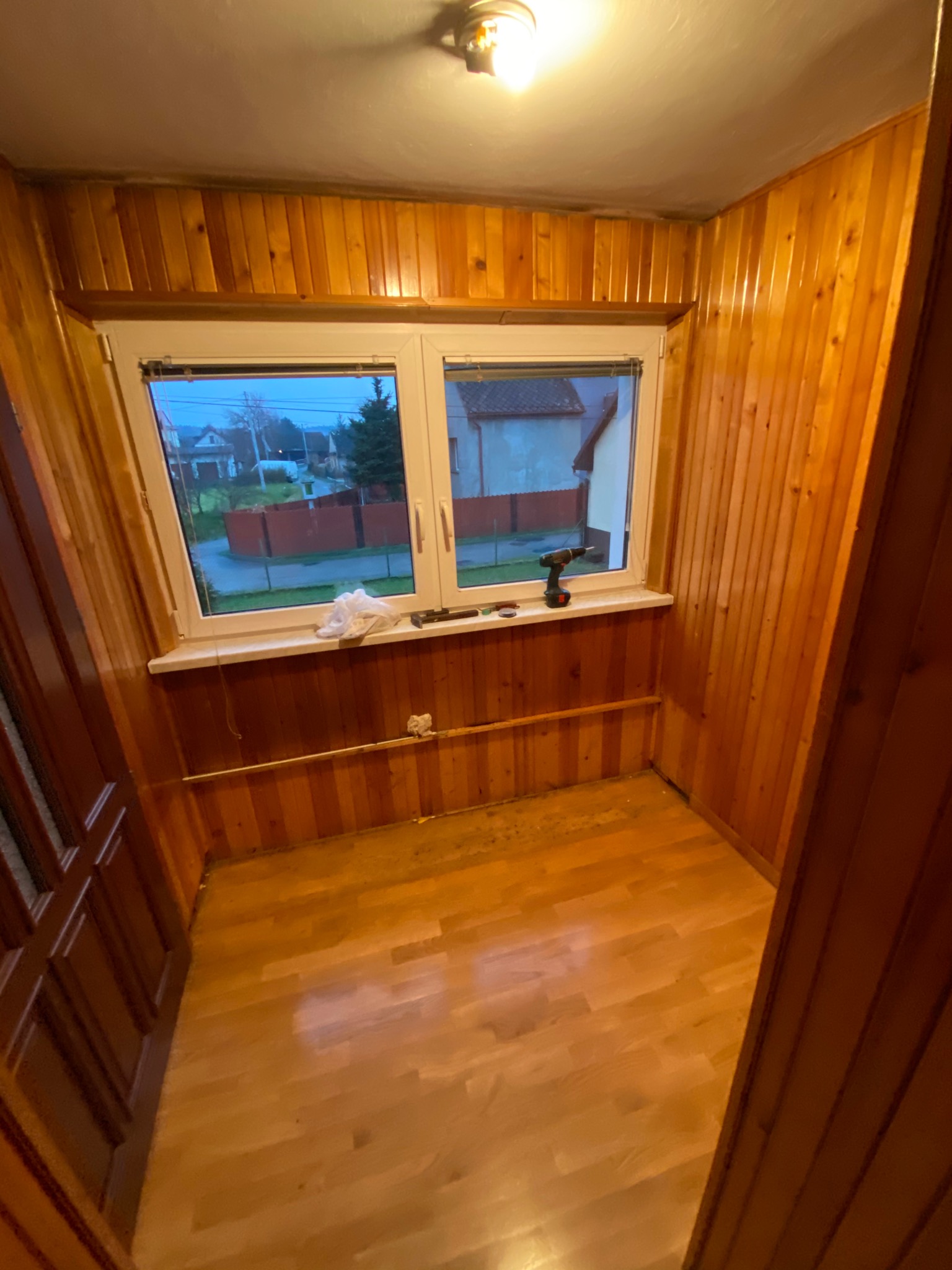 Wnętrze małego pomieszczenia wykończonego boazerią, z widocznym oknem, wiertarką na parapecie i fragmentem podłogi z desek.