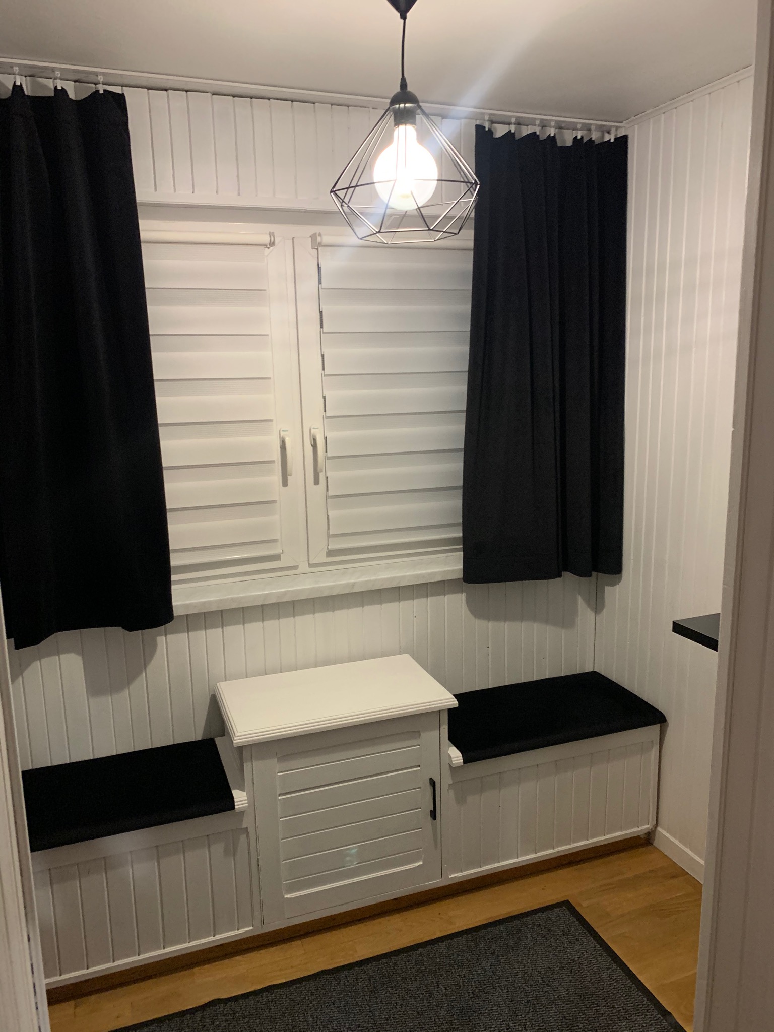 Białe panele ścienne z wbudowaną szafką i siedziskami, okno z żaluzjami, czarne zasłony i geometryczna lampa wisząca.