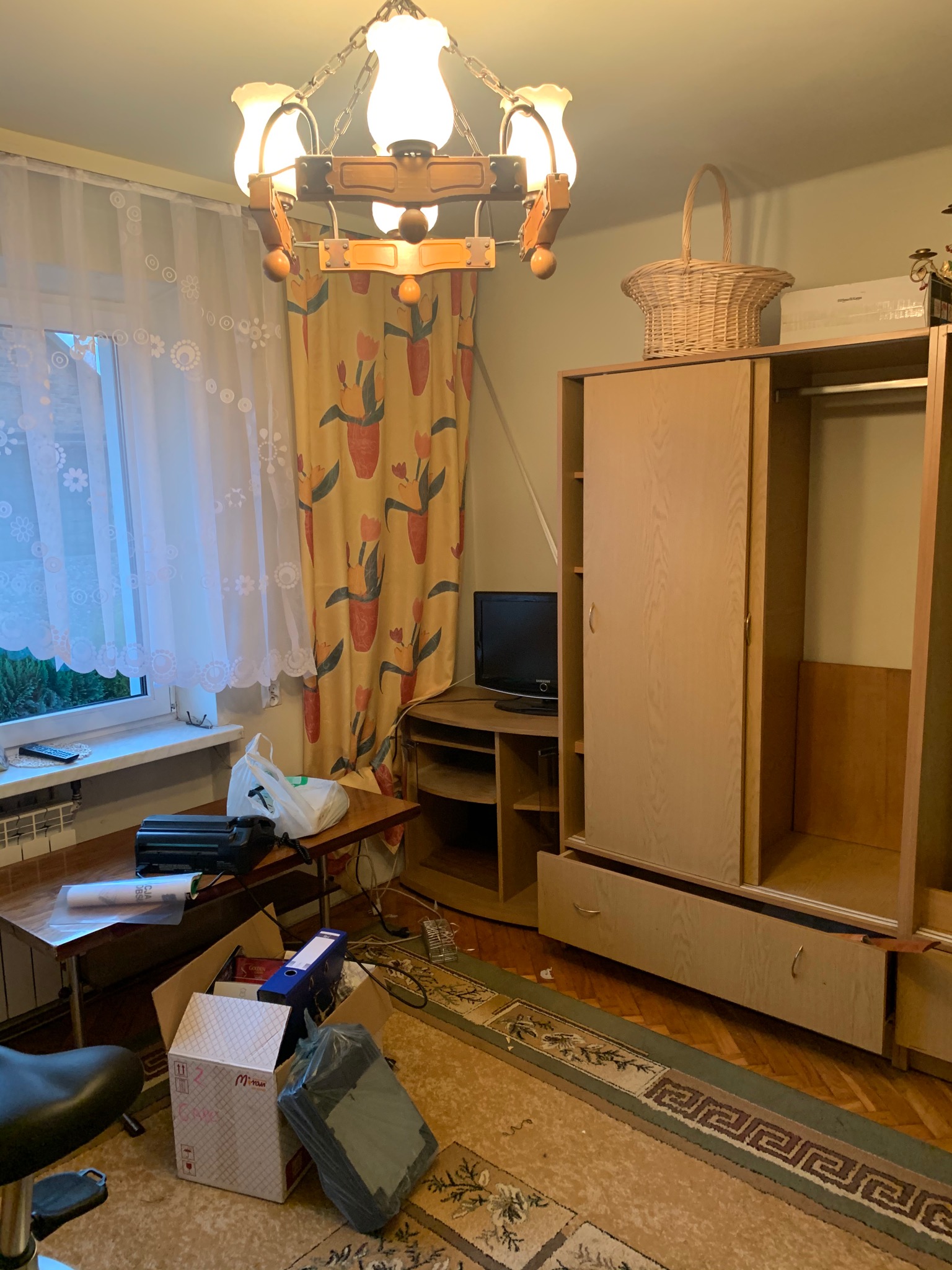 Pokój przed malowaniem: widoczny bałagan, meble odsunięte od ścian, kartony z rzeczami, przygotowania do malowania, lampa wisząca i zasłony w kwiaty.