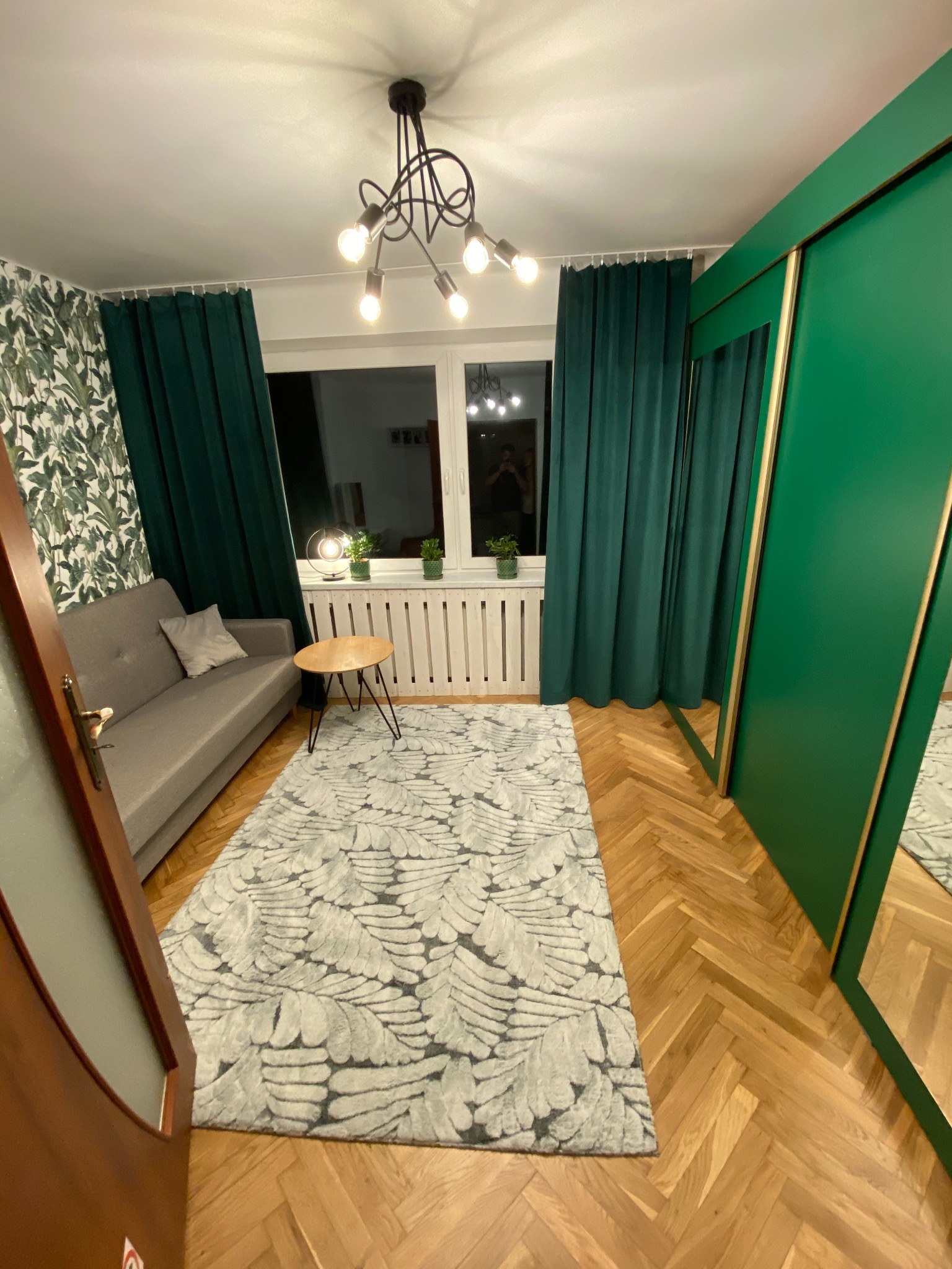 Salon w odcieniach zieleni i szarości: sofa, wzorzysty dywan, zielone zasłony i szafa przesuwna z lustrami, parkiet w jodełkę.