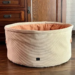 Okrągłe legowisko dla zwierząt z beżowego sztruksu z pomarańczowym wnętrzem, wykonane w szwalni w Opolu Lubelskim, stoi na drewnianej podłodze na tle drewnianej komody i drzwi.