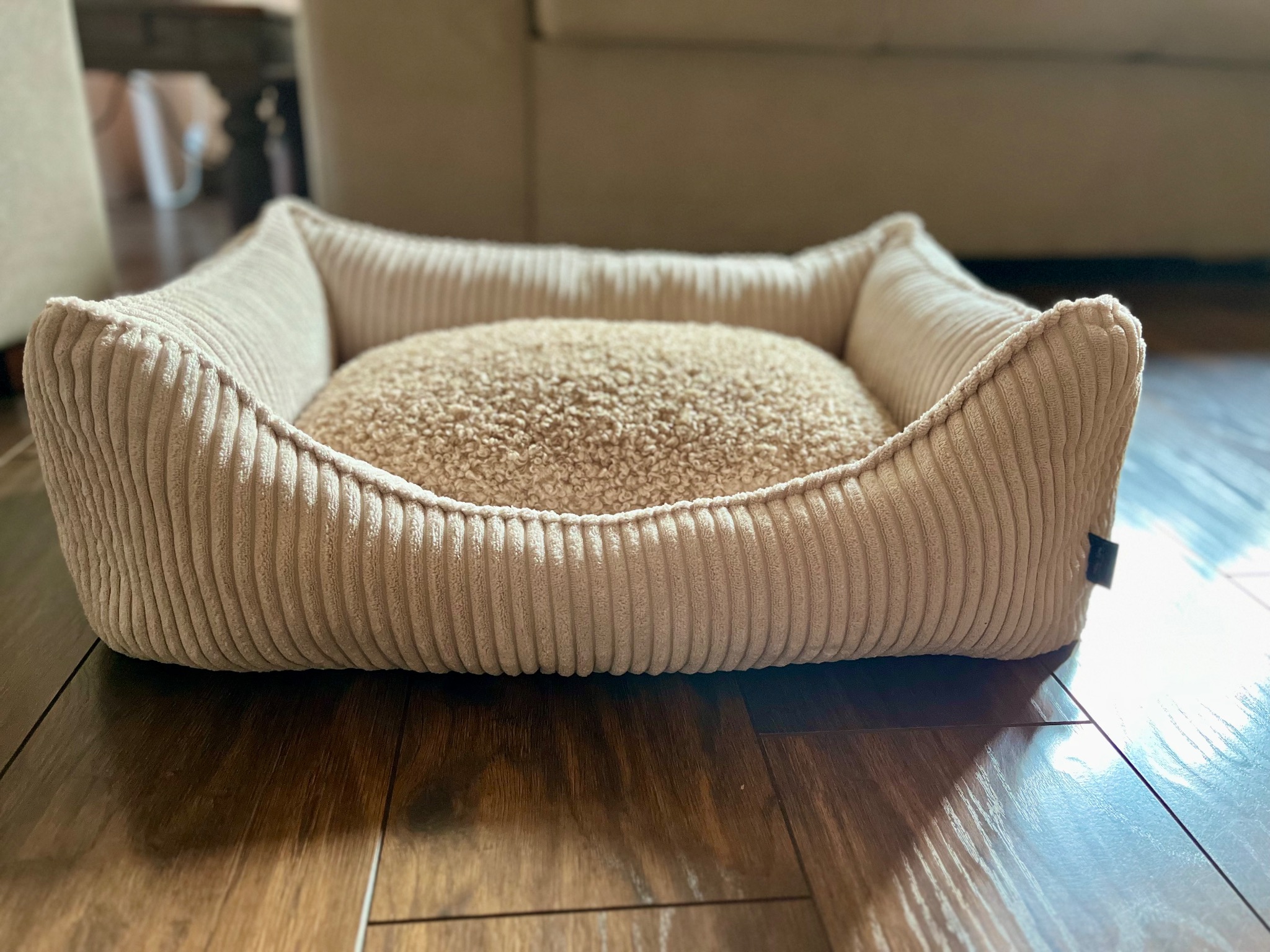 Beżowe, pluszowe legowisko dla psa z prążkowanej tkaniny na drewnianej podłodze w słonecznym pokoju.