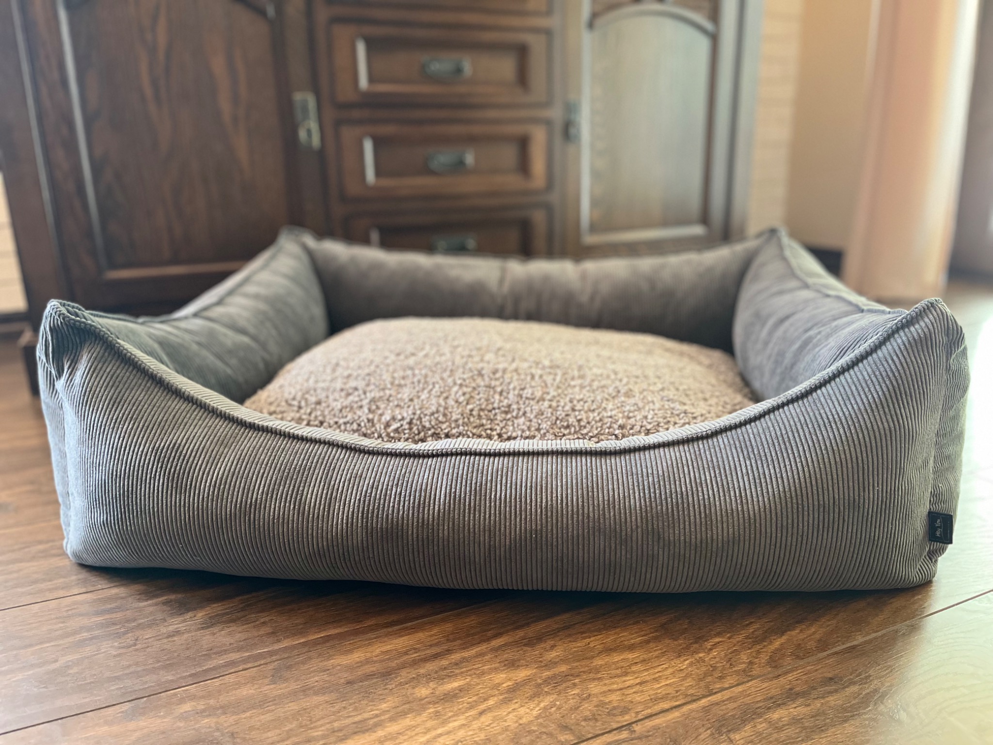 Szare, prążkowane legowisko dla psa z miękkim, beżowym wnętrzem, ustawione na drewnianej podłodze na tle ciemnych, drewnianych mebli.