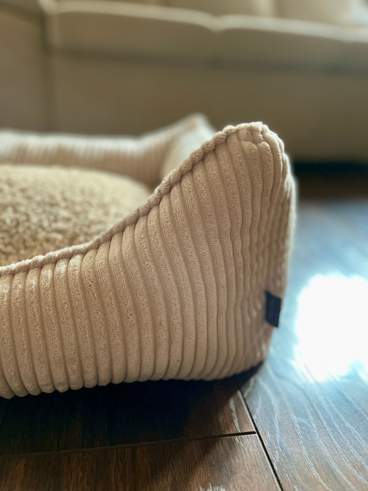 Beżowe posłanie dla psa z prążkowanej tkaniny na drewnianej podłodze, widoczny narożnik z małą metką.