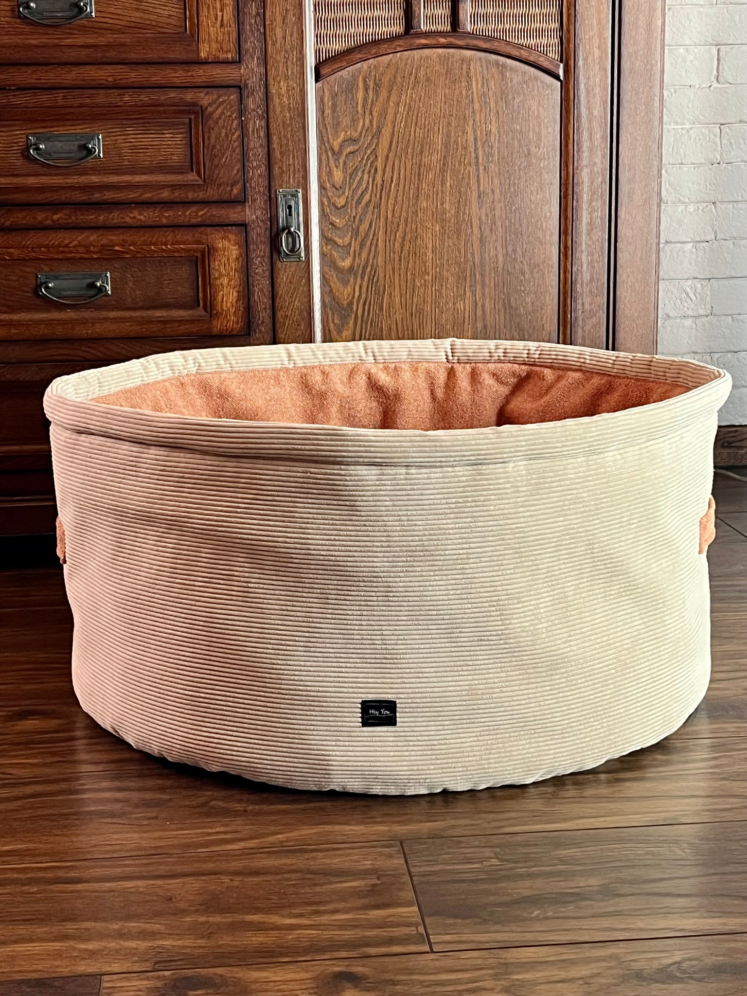 Okrągłe legowisko dla zwierząt z beżowego sztruksu z pomarańczowym wnętrzem, wykonane w szwalni w Opolu Lubelskim, stoi na drewnianej podłodze na tle drewnianej komody i drzwi.
