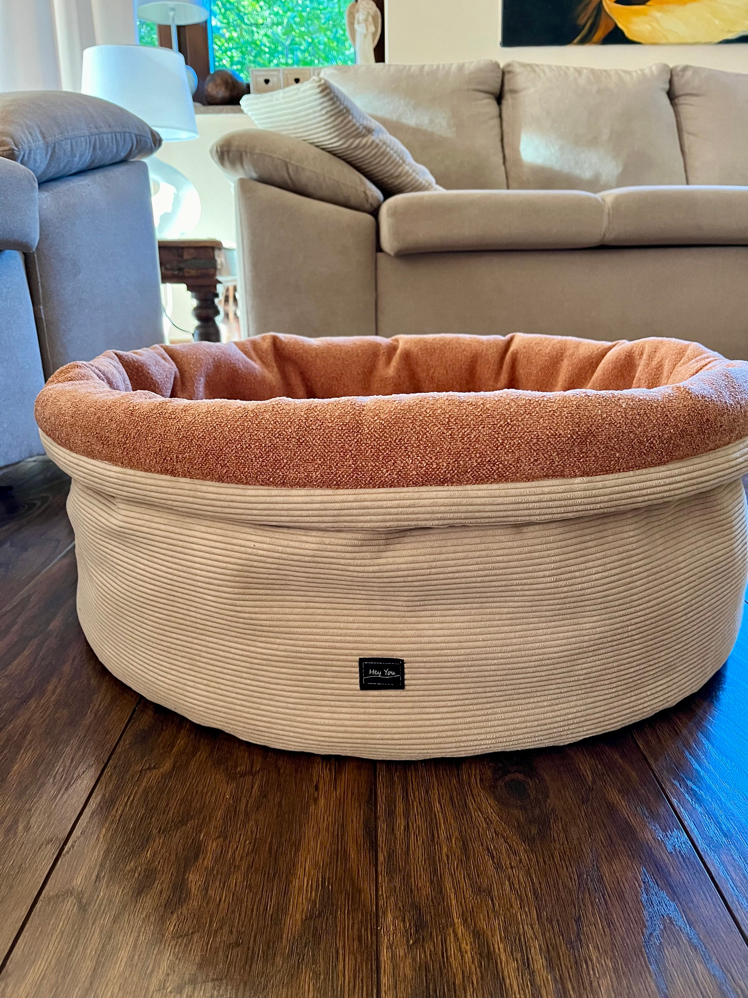 Okrągłe legowisko dla zwierząt z beżowego sztruksu i pomarańczowego materiału wewnątrz, umieszczone na drewnianej podłodze w salonie z sofą w tle.