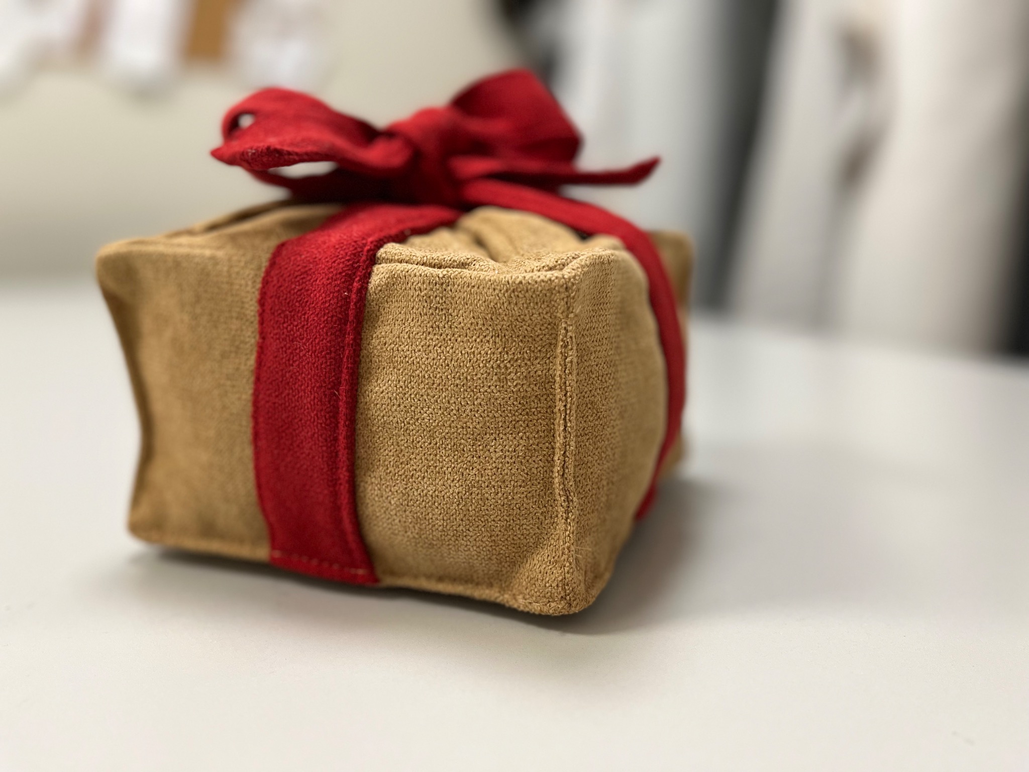 Beżowy, kwadratowy pokrowiec z grubej tkaniny, przewiązany czerwoną wstążką z kokardą, leży na białym blacie. Widoczne szwy i faktura materiału.