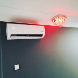 Eldar-Ice-Trans Adrian Folusiak - Biały klimatyzator ścienny z wyświetlaczem LCD na ciemnoszarej ścianie w pokoju z ozdobnym, czerwonym oświetleniem sufitowym