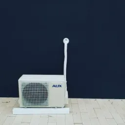 Biała jednostka zewnętrzna klimatyzatora marki AUX na drewnianej podłodze, na tle ciemnej ściany.