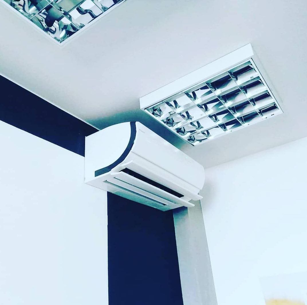 Biały klimatyzator ścienny z otwartą klapą w pomieszczeniu biurowym z czarno-białymi ścianami i lampami sufitowymi.