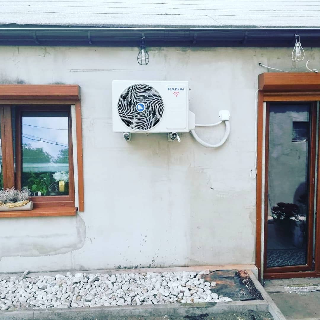 Biała jednostka zewnętrzna klimatyzatora marki Kaisai zamontowana na ścianie budynku z oknem i drzwiami z drewnianą stolarką, widoczna instalacja elektryczna i rury chłodnicze.