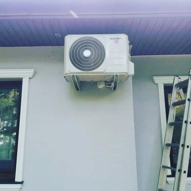 Biała jednostka zewnętrzna klimatyzatora marki Kaisai zamontowana na elewacji budynku, widoczna drabina oparta o ścianę.