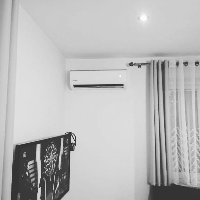 Czarno-białe zdjęcie wnętrza pokoju z zamontowanym klimatyzatorem ściennym, widoczne zasłony, plakat z słuchawkami na ścianie i włączone oświetlenie sufitowe.