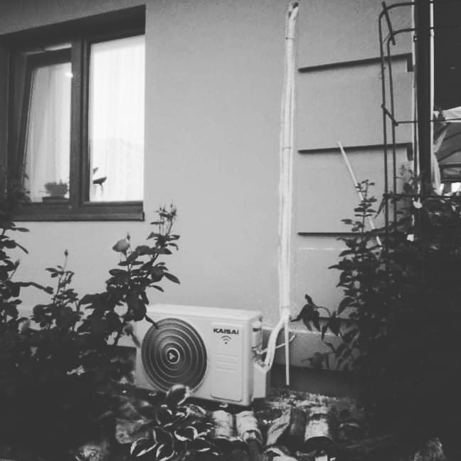 Zewnętrzna jednostka klimatyzacji Kaisai zamontowana przy ścianie domu, widoczna zza roślinności w czarno-białej tonacji, z rurami biegnącymi wzdłuż elewacji.