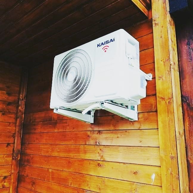Biała jednostka zewnętrzna klimatyzacji marki Kaisai zamontowana na drewnianej ścianie domu z boazerii.