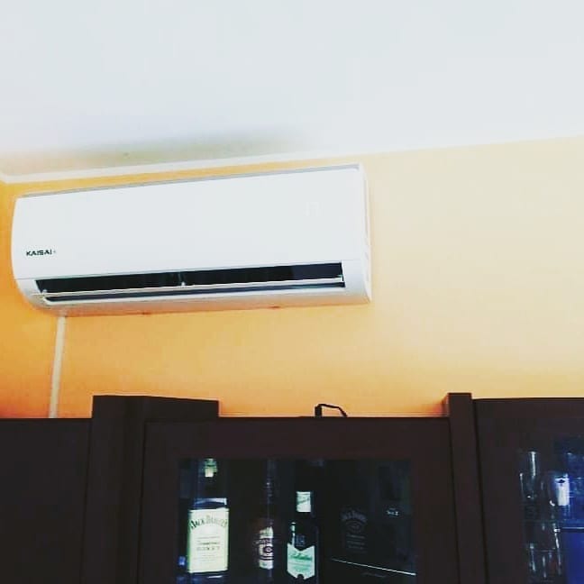 Biała jednostka klimatyzacyjna Kaisai zamontowana na pomarańczowej ścianie nad ciemnym barkiem z butelkami alkoholu.
