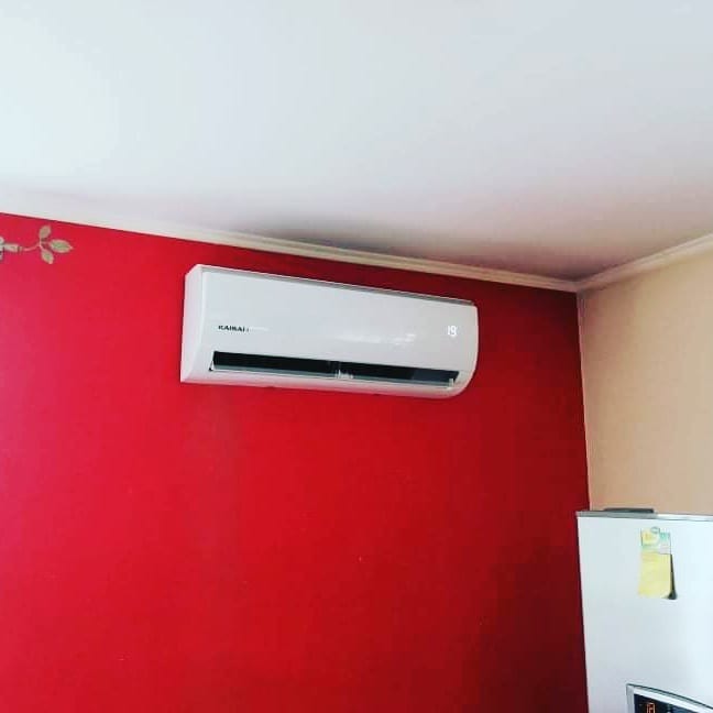 Biały klimatyzator marki Kaisai zamontowany na czerwonej ścianie w pomieszczeniu, obok widoczna lodówka