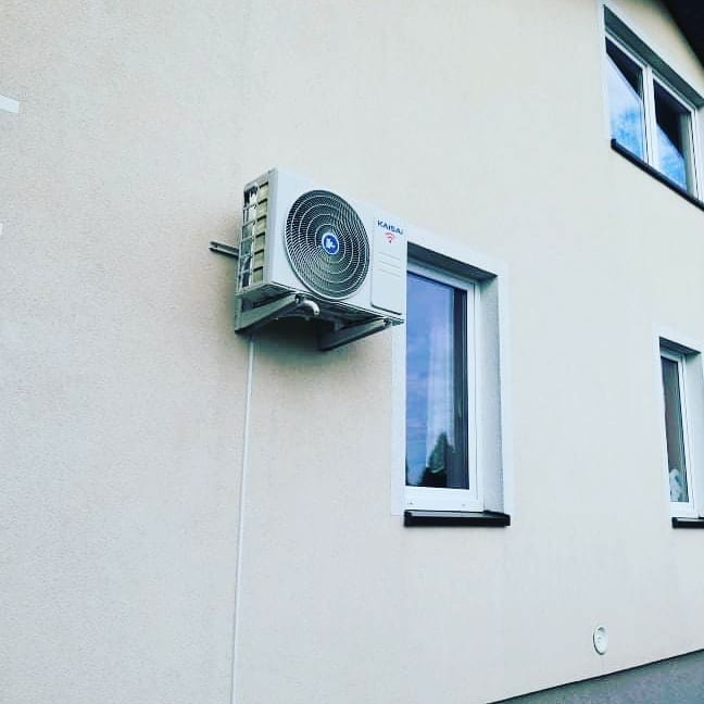 Zewnętrzna jednostka klimatyzacji marki Kaisai, zamontowana na elewacji budynku mieszkalnego obok okna z widocznym odbiciem nieba.