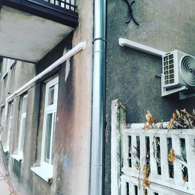 Zewnętrzna jednostka klimatyzacji zamontowana na szarej, zewnętrznej ścianie budynku obok rynny spustowej, widoczna zza starego betonowego ogrodzenia z uschniętymi liśćmi.