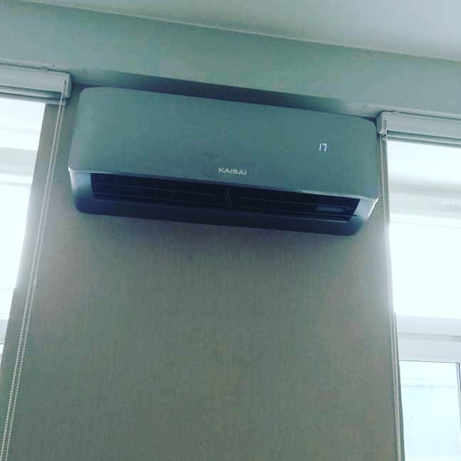 Szary klimatyzator Kaisai zamontowany na ścianie, wyświetlający temperaturę 17 stopni, obok okna z roletą.