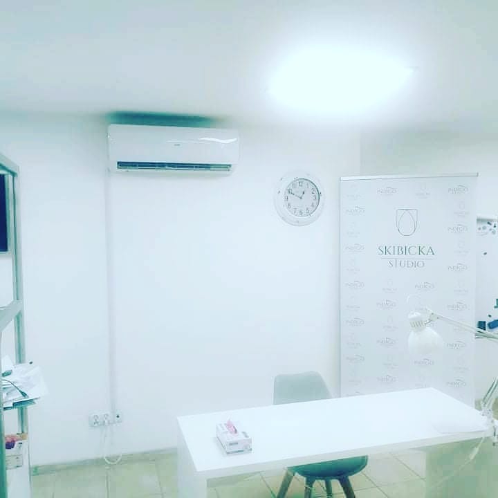 Wnętrze gabinetu kosmetycznego z zamontowanym klimatyzatorem ściennym, zegarem, biurkiem i roll-upem reklamowym.