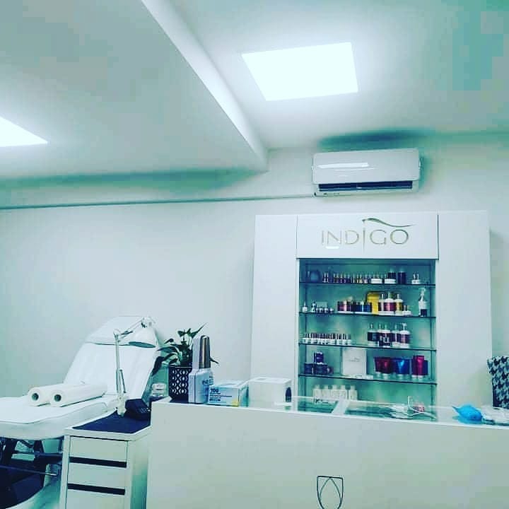 Jasne wnętrze salonu kosmetycznego z klimatyzatorem ściennym, regałem z produktami Indigo i białym fotelem zabiegowym.