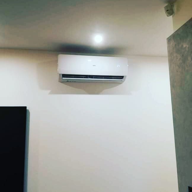 Biała klimatyzacja ścienna zamontowana w pomieszczeniu z białymi ścianami, widoczny czujnik ruchu na suficie i fragment szarej ściany po prawej stronie.