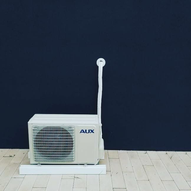 Biała jednostka zewnętrzna klimatyzatora marki AUX na drewnianej podłodze, na tle ciemnej ściany.