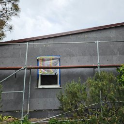 Krystian Porwol Usługi Budowlane - Dom w trakcie prac, ściana pokryta szarym styropianem, okno zabezpieczone folią, widoczne rusztowanie i roślinność.