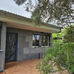 Krystian Porwol Usługi Budowlane - Budynek w trakcie prac związanych z izolacją termiczną, widoczne szare płyty izolacyjne na elewacji, okno zabezpieczone taśmą, a drzwi folią ochronną, na pierwszym planie taras z terakoty i zieleń.