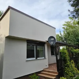 Ocieplona elewacja domu jednorodzinnego z brązowym orynnowaniem i anteną satelitarną, widok z boku na wejście z drewnianymi schodami i pergolą porośniętą zielenią.