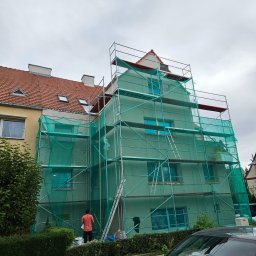 Krystian Porwol Usługi Budowlane - Budynek w trakcie prac elewacyjnych, osłonięty siatką ochronną na rusztowaniu. Widoczny pracownik w pomarańczowej koszulce i wiadro z farbą. Ujęcie z dołu, pochmurne niebo.