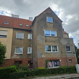 Krystian Porwol Usługi Budowlane - Dwupiętrowy budynek z widocznymi śladami uszkodzeń elewacji, z czerwonym dachem i oknami. Na parterze okna z reklamami. Osoba przy budynku. Pochmurne niebo.