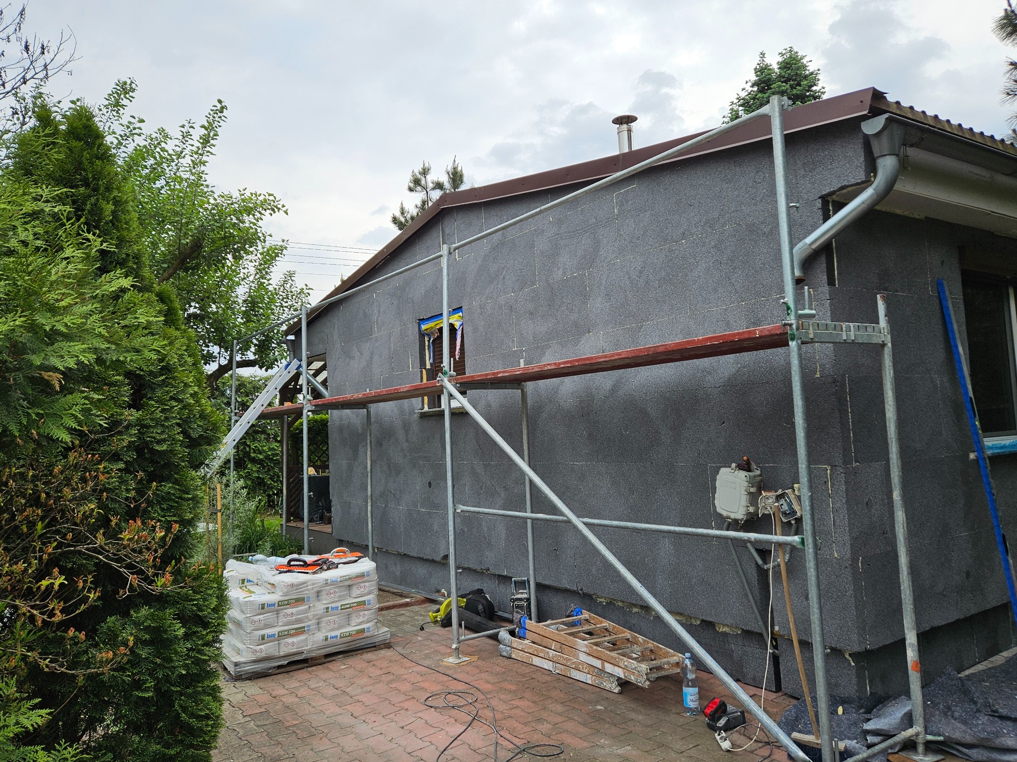 Dom jednorodzinny w trakcie prac związanych z ociepleniem elewacji, widoczne rusztowanie, płyty styropianowe w kolorze grafitowym, paleta z workami kleju i narzędzia na brukowanym podłożu.