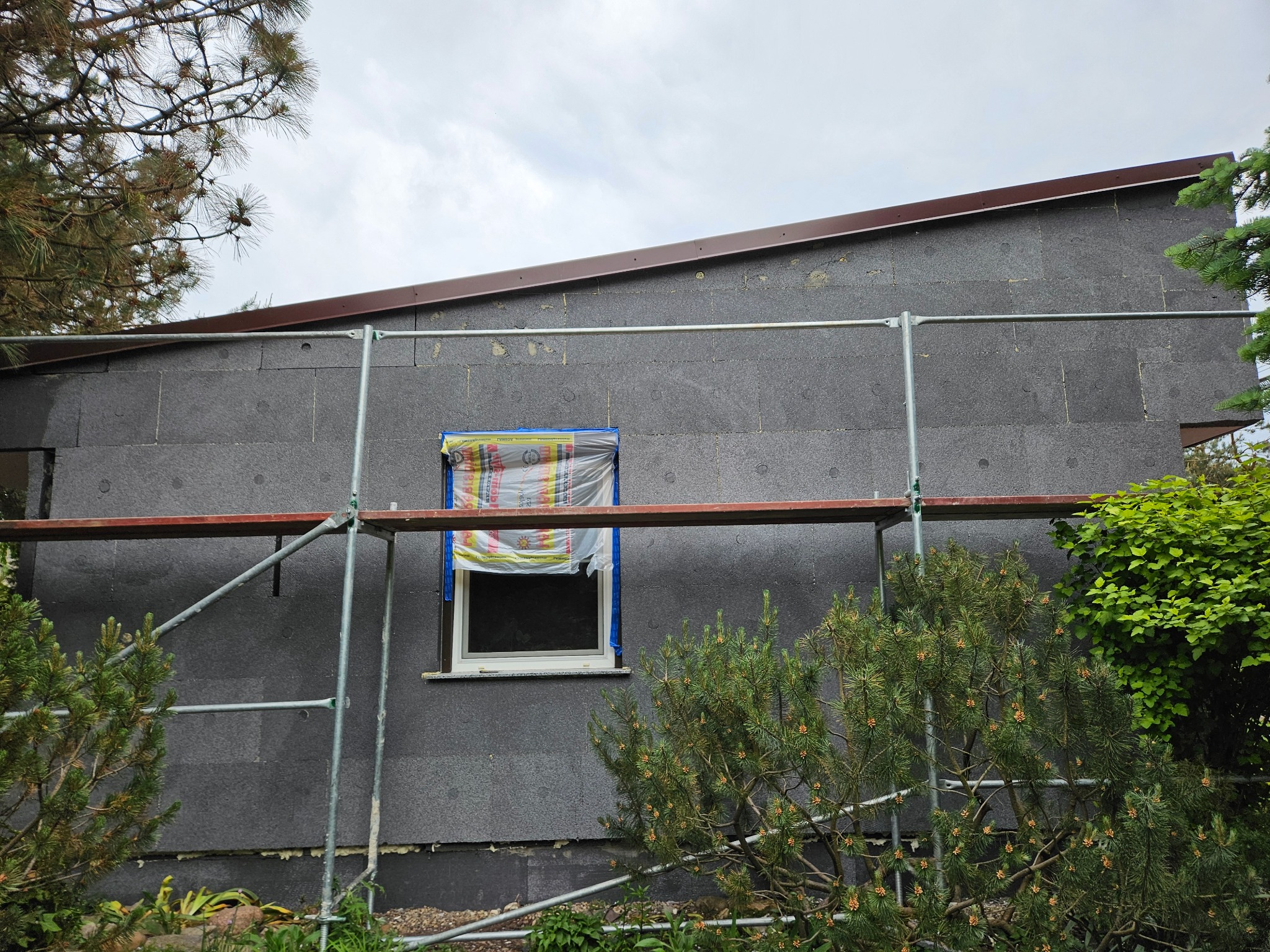 Dom w trakcie prac, ściana pokryta szarym styropianem, okno zabezpieczone folią, widoczne rusztowanie i roślinność.